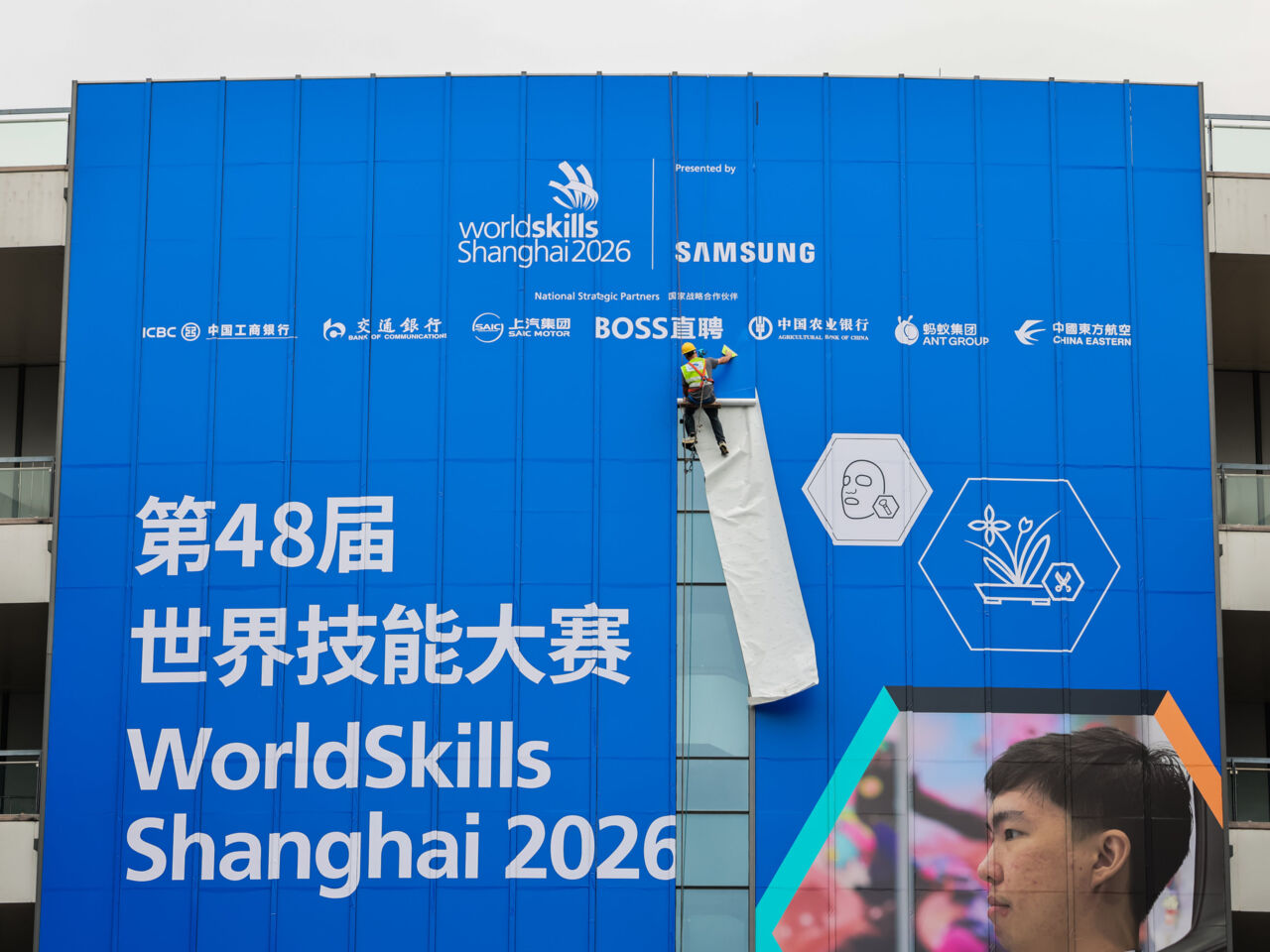 A construction worker climbing down a massive sign advertising WorldSkills Shanghai 2026.